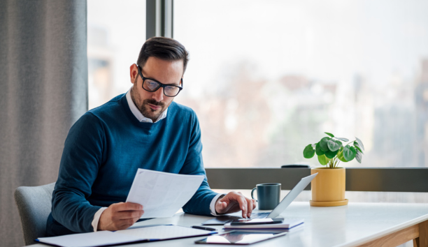 Man in blue sweater with glasses looks down at paper in his hand while sitting at a desk working. 